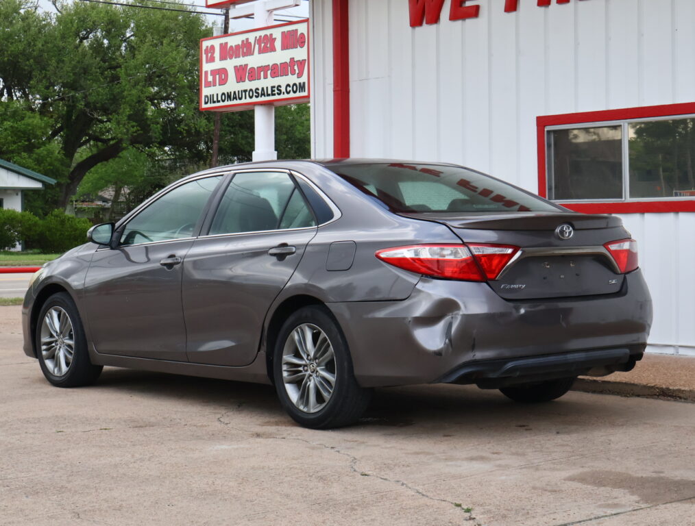 Rear three-quarter view of a gray Toyota Camry SE with a dented rear bumper in a car dealership lot.