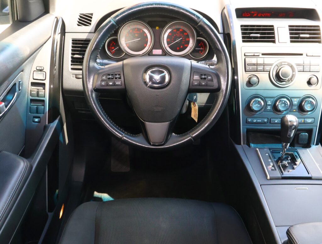 Car interior focusing on the steering wheel with Mazda emblem and the instrument cluster in view across the dashboard center stack.