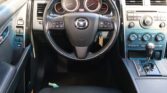 Car interior focusing on the steering wheel with Mazda emblem and the instrument cluster in view across the dashboard center stack.