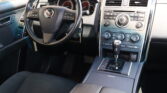 Front interior of a car: steering wheel with Mazda logo, instrument cluster behind, and center console with climate controls and radio.