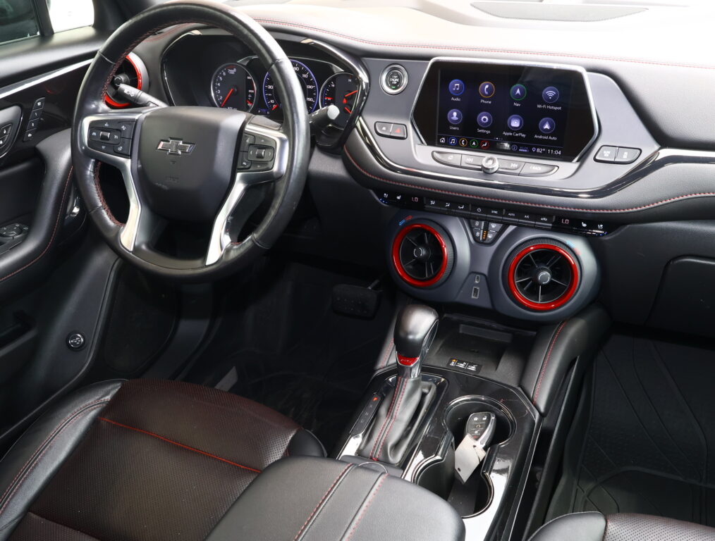 Front interior of a black car showing leather steering wheel with red stitching, digital gauges, and a large touchscreen infotainment display.
