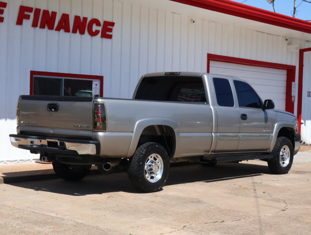 Side view of a silver Chevrolet Silverado pickup truck parked in front of a white building with red trim and a large red 'FINANCE' sign partially visible.