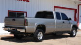 Side view of a silver Chevrolet Silverado pickup truck parked in front of a white building with red trim and a large red 'FINANCE' sign partially visible.