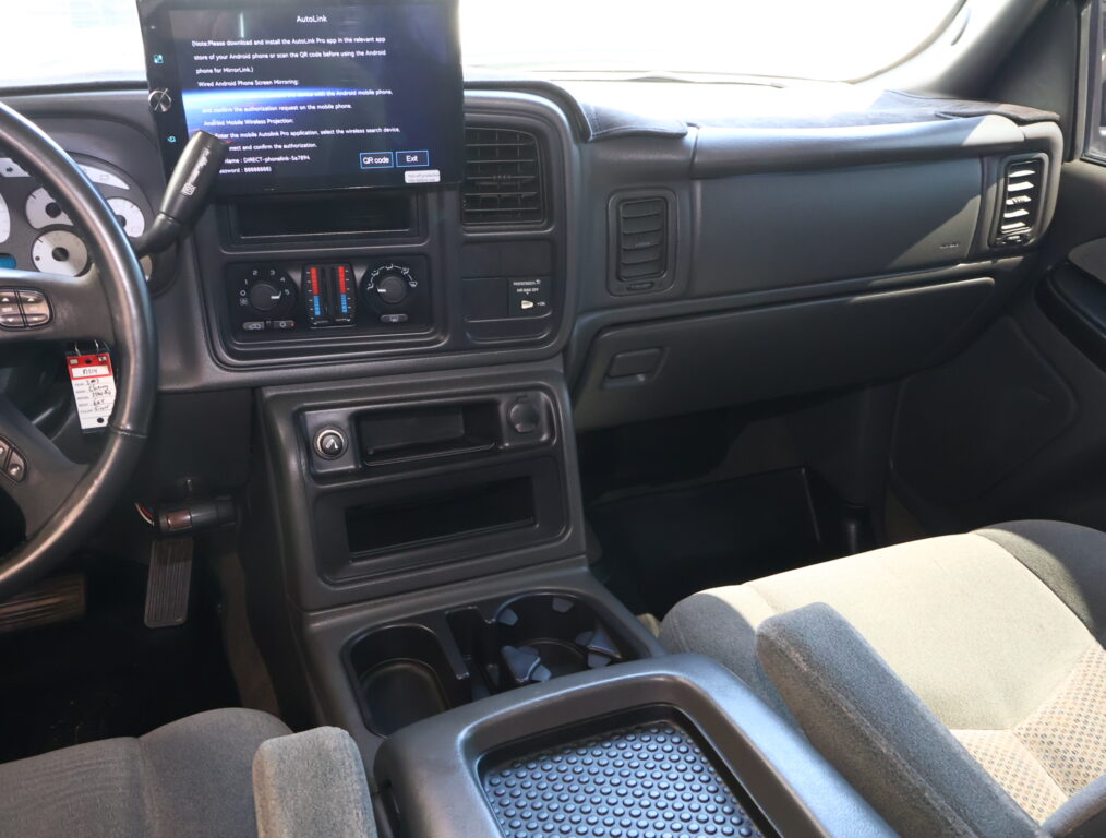 Interior of a vehicle cabin focused on the driver's side: steering wheel, dashboard with a mounted tablet screen, climate controls, and cupholders between the seats.