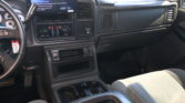 Interior of a vehicle cabin focused on the driver's side: steering wheel, dashboard with a mounted tablet screen, climate controls, and cupholders between the seats.