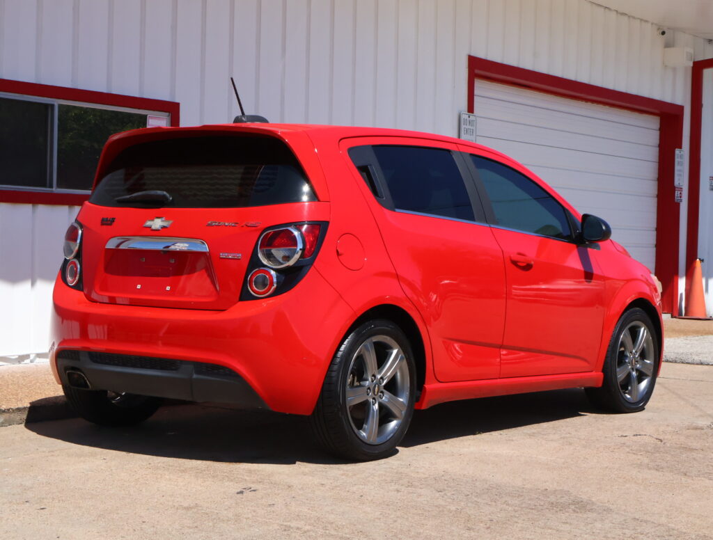 Red Chevrolet Sonic RS parked near a white building with a large garage door, seen from the rear-side angle