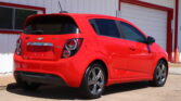 Red Chevrolet Sonic RS parked near a white building with a large garage door, seen from the rear-side angle