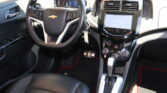 Black Chevrolet car interior focusing on the steering wheel, digital instrument cluster, and glossy center infotainment screen with gear shifter nearby