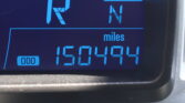 Blue backlit car instrument panel displaying compass indicator with gear positions R and N and an odometer label measuring miles (informational image).