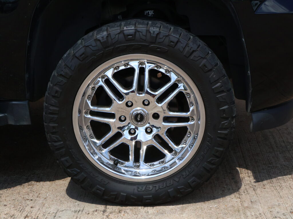 Close-up of a chrome alloy wheel and large all-terrain tire mounted on a dark vehicle, wheel filling the frame.