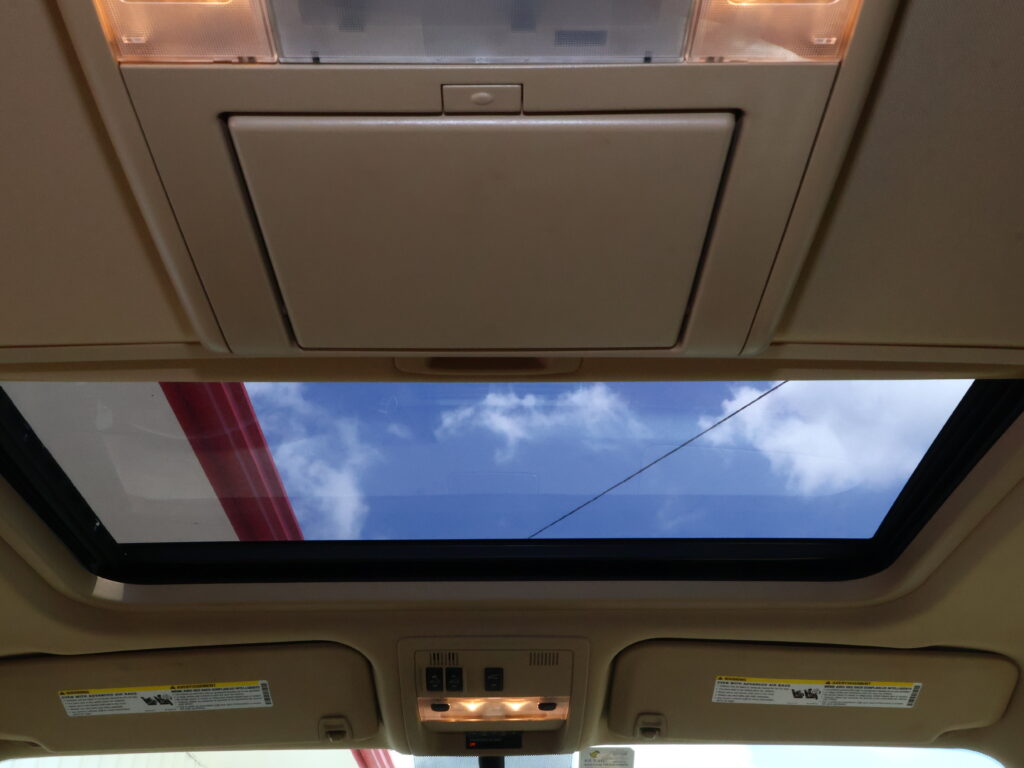 View of a vehicle interior ceiling with an open sunroof, showing blue sky and clouds through the glass, and overhead lights nearby