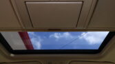 View of a vehicle interior ceiling with an open sunroof, showing blue sky and clouds through the glass, and overhead lights nearby
