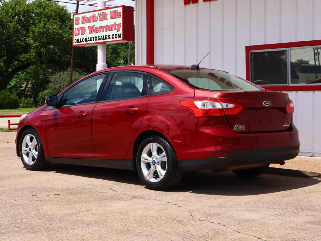 Red Ford Focus parked in a lot beside a white building with a large window; sign advertises a 12‑month/12k mile warranty.