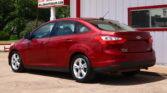 Red Ford Focus parked in a lot beside a white building with a large window; sign advertises a 12‑month/12k mile warranty.