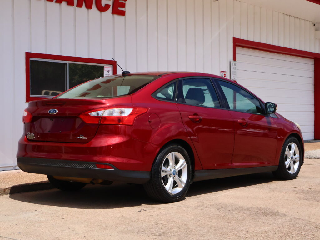 Red Ford Focus sedan parked in front of a white building with red trim and a rolling door visible in the background, seen from the rear side of the car.