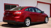 Red Ford Focus sedan parked in front of a white building with red trim and a rolling door visible in the background, seen from the rear side of the car.