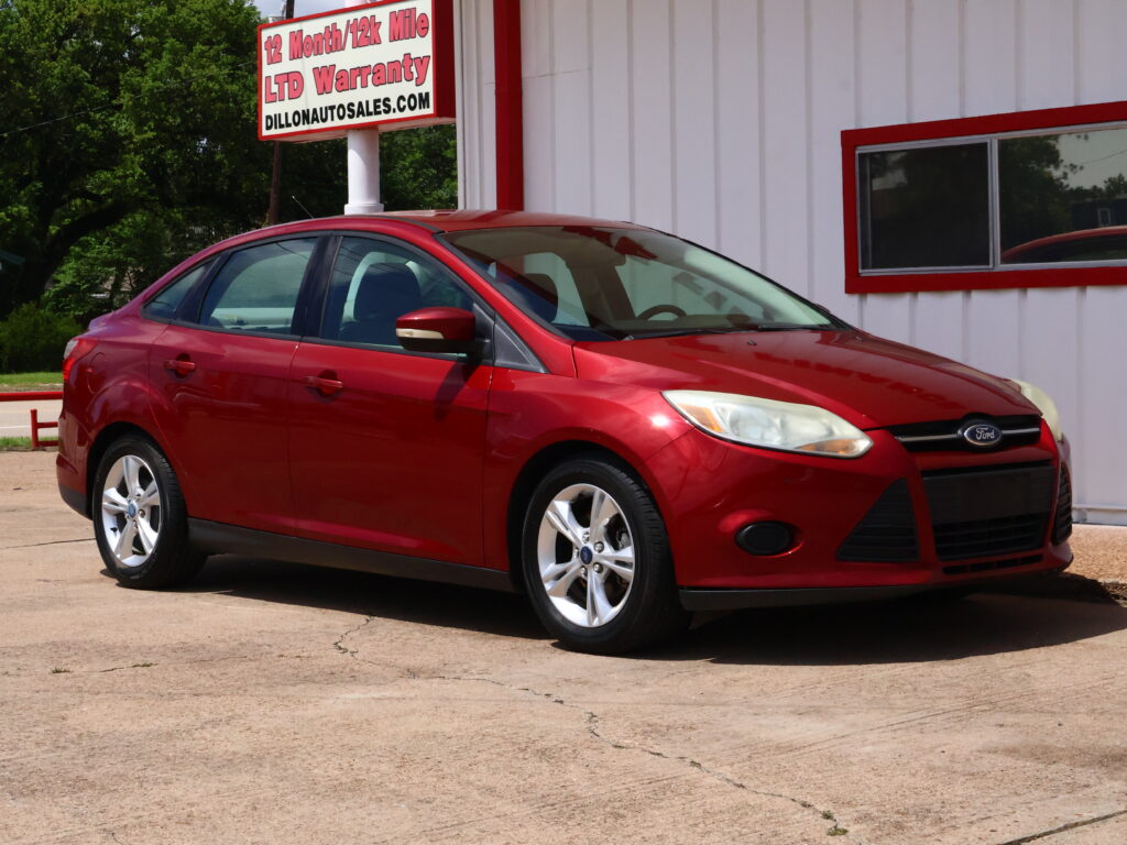 Red Ford sedan parked on a concrete lot beside a white building with red trim and a large sign advertising a 12‑month/12k‑mile warranty in the background.