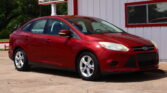 Red Ford sedan parked on a concrete lot beside a white building with red trim and a large sign advertising a 12‑month/12k‑mile warranty in the background.