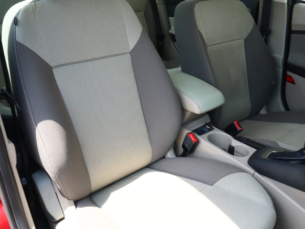 Car interior showing two front seats with light gray fabric and a center armrest between them, plus visible seat belts and cupholders.
