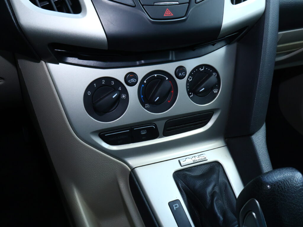 Center console of a car showing climate control dials, hazard button, and a gear selector area; includes A/C and defrost controls and a 'Powered by Microsoft' badge on the panel below.