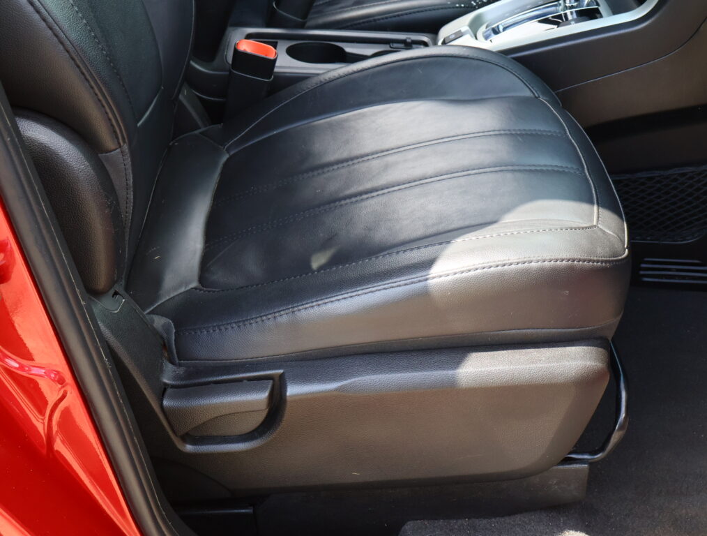 Close-up of a black leather car seat with the center console and orange-seat-belt buckle nearby, red door frame visible at left.