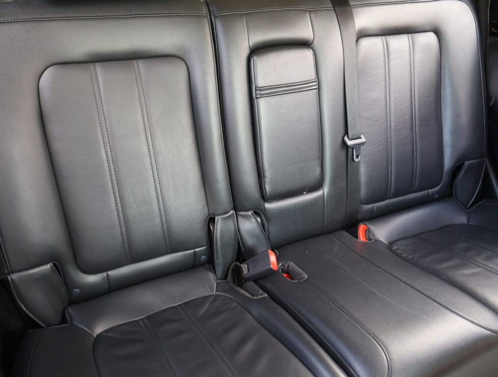 Leather car back seat with two visible seat belts and red buckles in a dark interior.