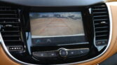 In-car touchscreen showing the rearview camera feed with orange parking guideline lines, surrounded by brown leather dashboard and air vents in a vehicle interior.