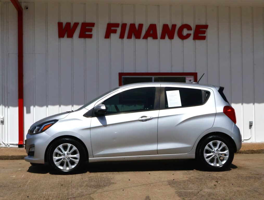 Silver hatchback parked in front of a white wall with a red 'WE FINANCE' sign above the car's roof line.