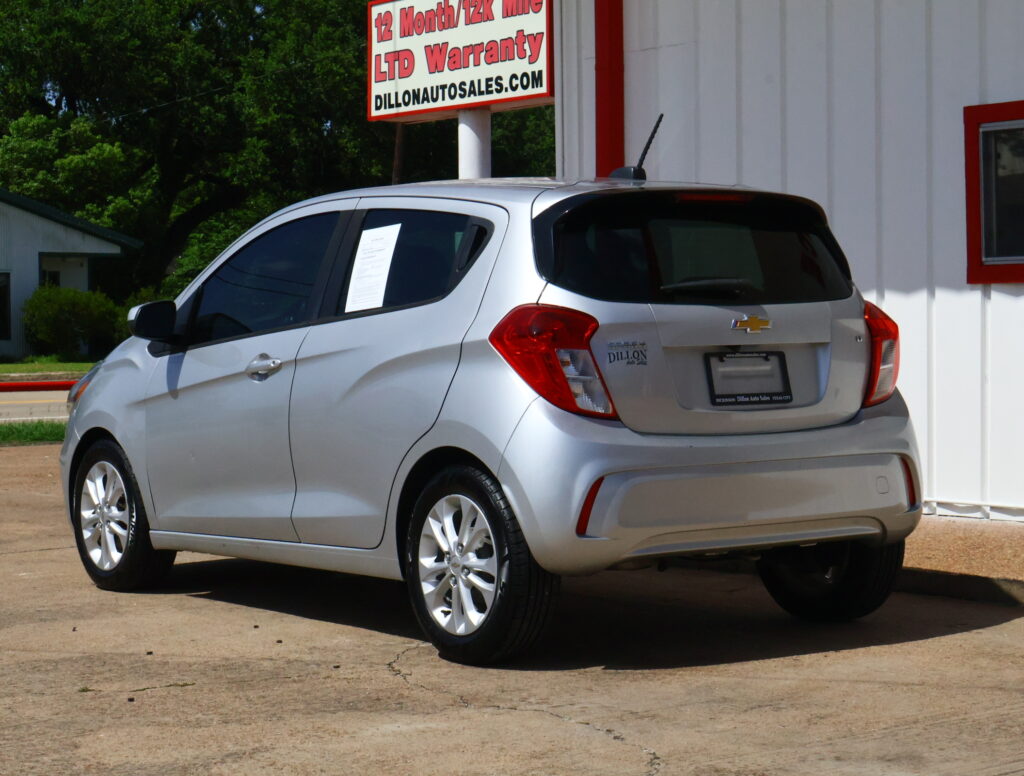 Rear three-quarter view of a silver Chevrolet SUV parked in a dealership lot, with a large sign offering a 12-month/12K-mile warranty above the building.