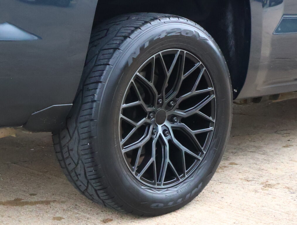 Close-up of a car tire and alloy wheel with dark, intricate spokes showing a low-angle perspective of the wheel and fender.