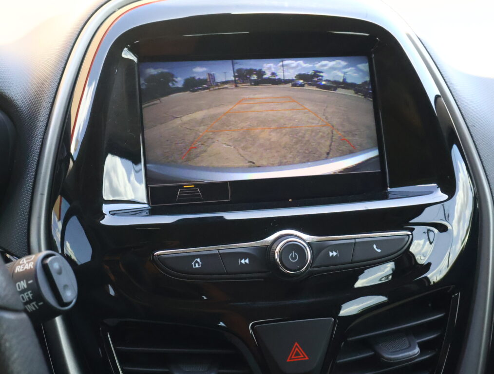 Display of a car's rearview camera with orange guiding lines over a parking lot scene, aiding reverse parking.