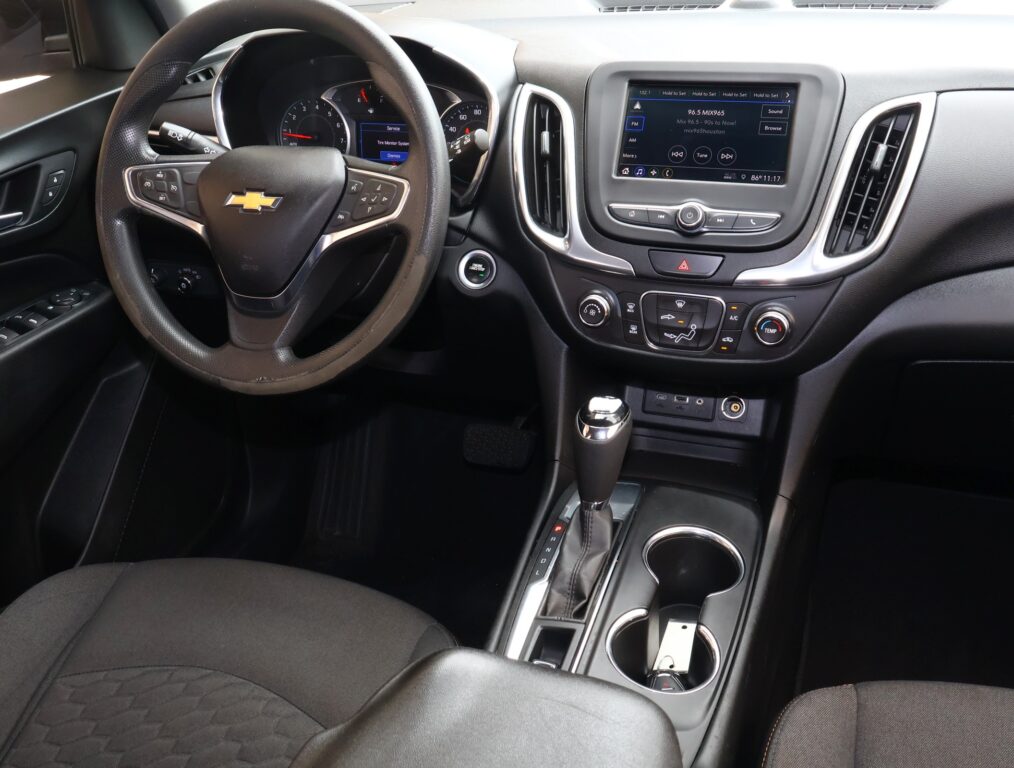 Interior view of a car cabin: steering wheel with Chevrolet logo, central touchscreen, gear shifter, and cup holders in a black dashboard area.