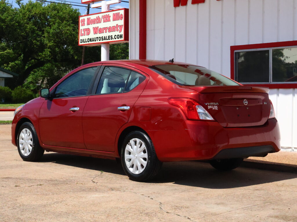 Red Nissan Versa sedan parked at a car dealership, white building with red trim in the background and a sign visible nearby.