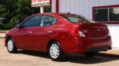 Red Nissan Versa sedan parked at a car dealership, white building with red trim in the background and a sign visible nearby.