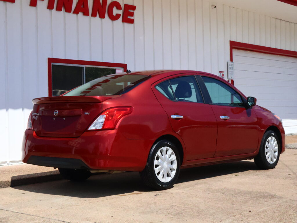 Red Nissan Versa parked in front of a white building with red trim and a partially visible 'FINANCE' sign, shown from the rear side.