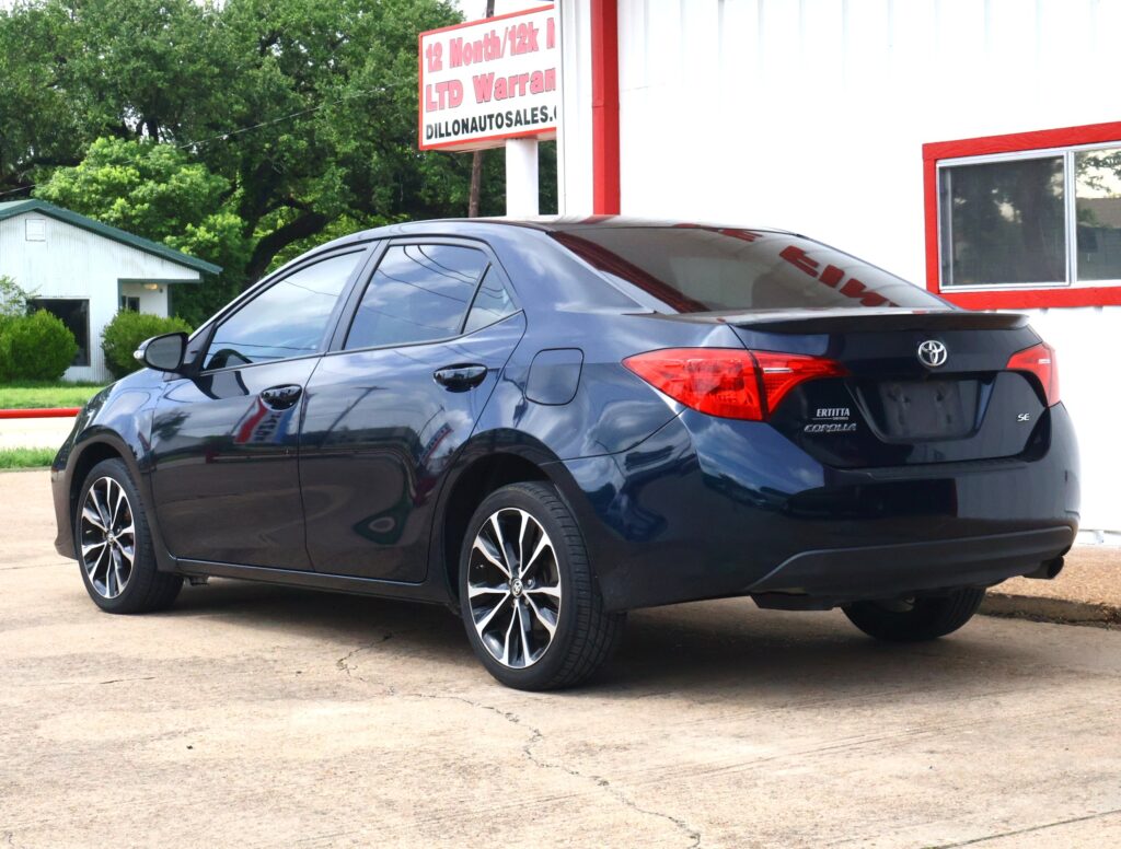 Rear three-quarter view of a dark blue Toyota Corolla parked on a lot beside a white building with red trim and a dealership sign in the background.