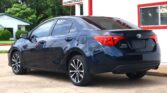 Rear three-quarter view of a dark blue Toyota Corolla parked on a lot beside a white building with red trim and a dealership sign in the background.