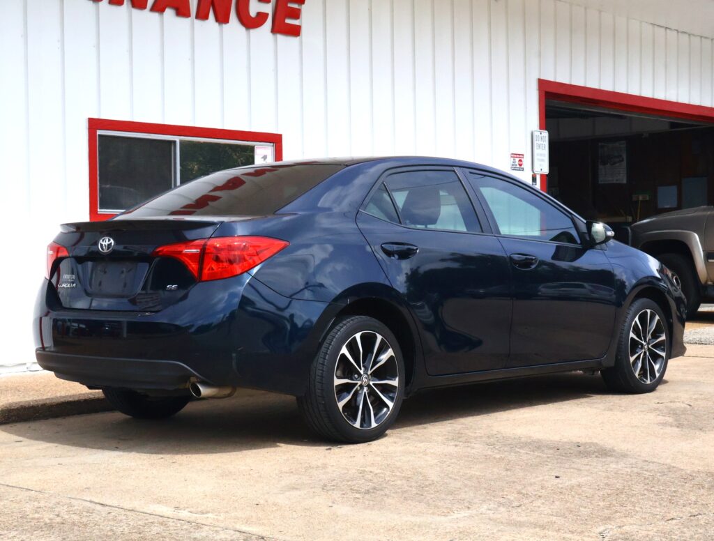 Rear three-quarter view of a dark blue Toyota Camry parked in front of a white building with red trim. A service entrance and a 'Do Not Enter' sign are visible to the right.
