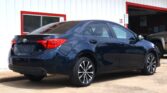 Rear three-quarter view of a dark blue Toyota Camry parked in front of a white building with red trim. A service entrance and a 'Do Not Enter' sign are visible to the right.