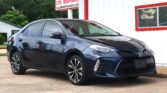 Dark gray Toyota sedan parked on a lot in front of a white, red-trimmed building with a sign offering 12‑month/12k‑mile warranty