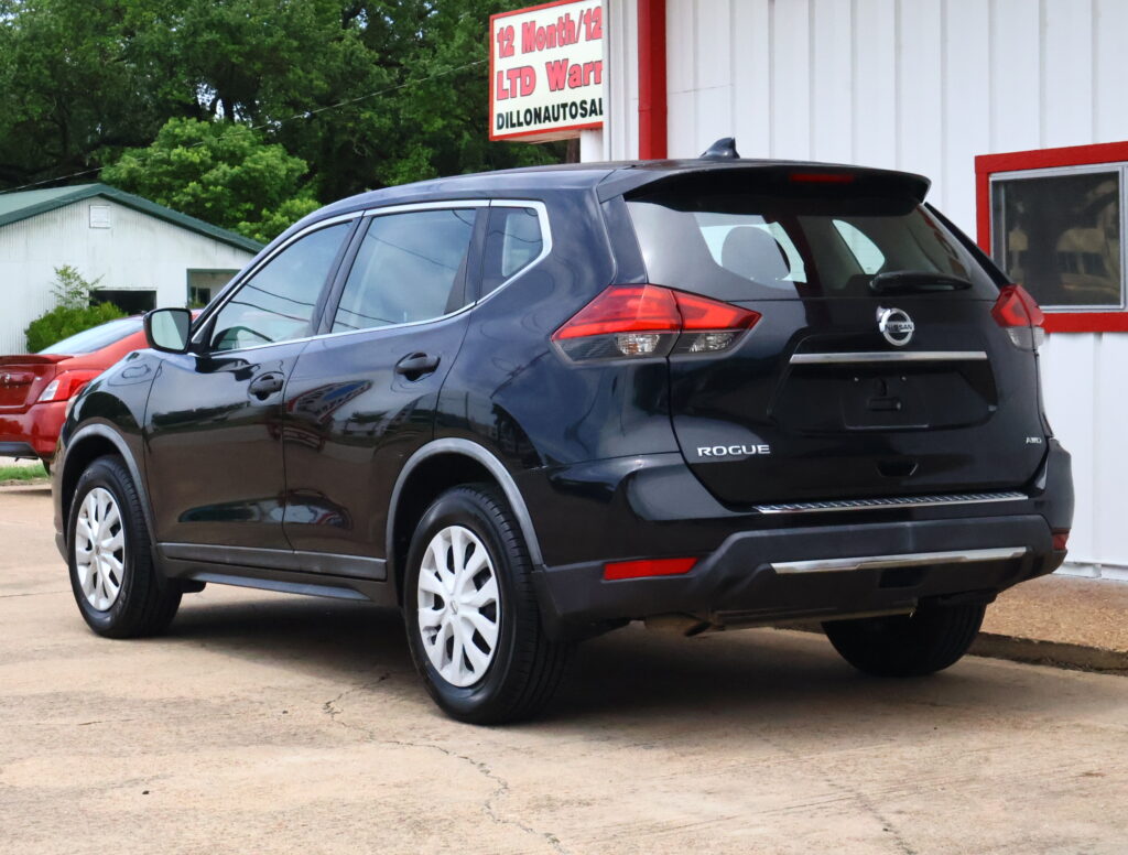 Rear three-quarter view of a dark gray Nissan Rogue SUV parked outside a white building with red trim and a sign above.