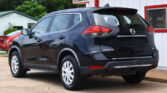 Rear three-quarter view of a dark gray Nissan Rogue SUV parked outside a white building with red trim and a sign above.