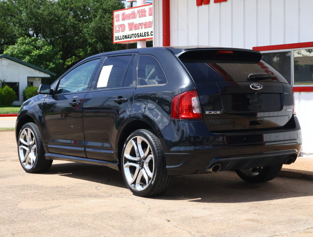 Black Ford SUV parked in front of a white building with red trim at a car lot, rear three-quarter view showing wheels and tailgate.