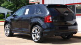 Black Ford SUV parked in front of a white building with red trim at a car lot, rear three-quarter view showing wheels and tailgate.