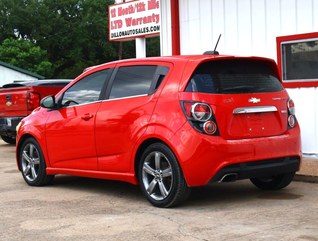 Red Chevrolet Sonic parked in front of a white building at a car dealership in daytime.