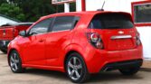 Red Chevrolet Sonic parked in front of a white building at a car dealership in daytime.