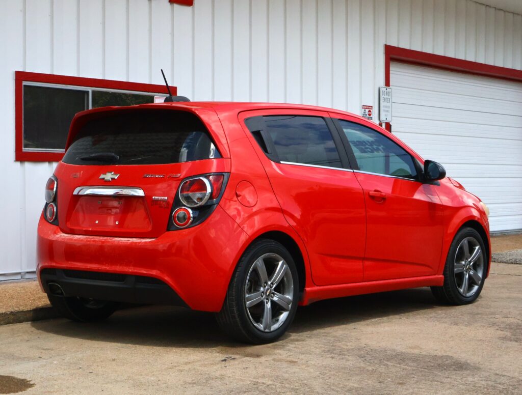 Rear three-quarter view of a bright red Chevrolet Sonic RS hatchback parked beside a white metal building with red trim and a small window.