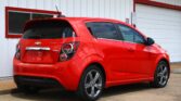 Rear three-quarter view of a bright red Chevrolet Sonic RS hatchback parked beside a white metal building with red trim and a small window.