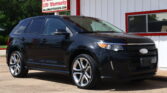 Black SUV parked in front of a white building with red trim and a sign advertising a 12-month/12k-mile LTD warranty at a car dealership.