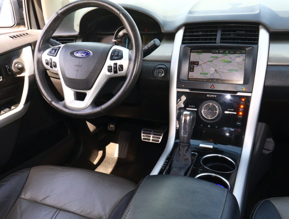 Driver’s seat view of a Ford interior: steering wheel with built‑in controls, dashboard gauges, and a center touchscreen displaying a navigation map alongside climate controls and gear selector in the foreground